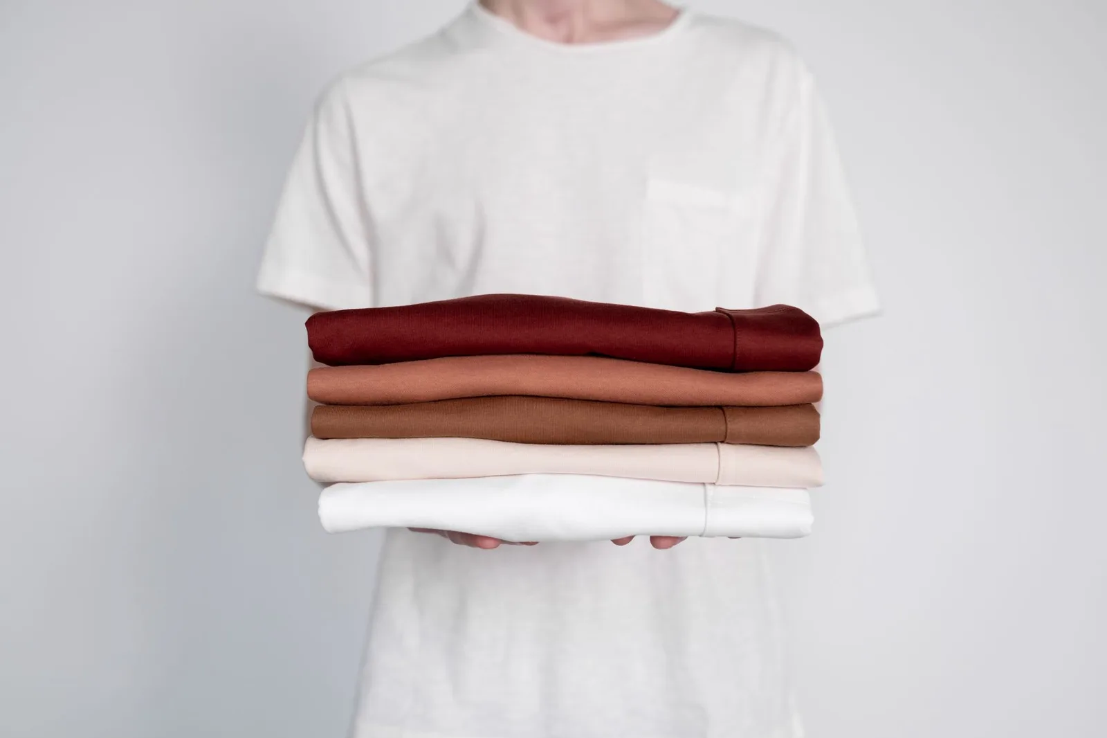 A stack of folded cotton T-shirts in neutral colors on a plain surface